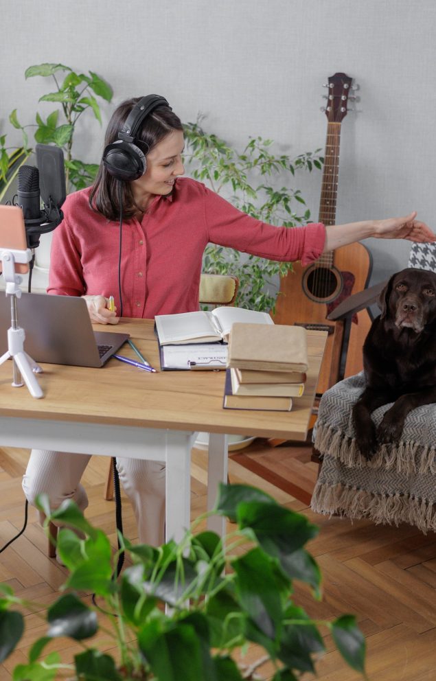 woman in a home office records podcast audio content with a microphone and headphones and a labrador